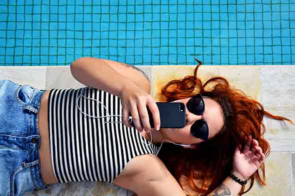 woman in sunglasses lying down beside pool with phone in hand