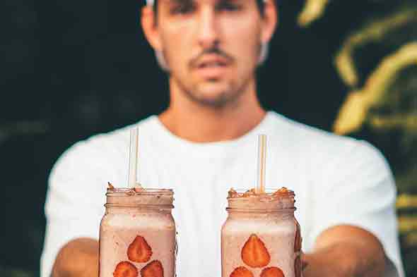 A man in a white shirt and cap holding two glasses of fruit shake